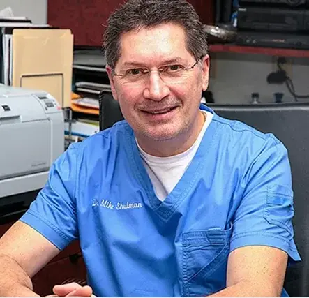 Dr. Mike Shulman, co-founder and dentist at R&S Dental Associates in Clifton, NJ, smiling in his office wearing blue scrubs.