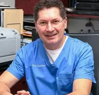 Dr. Mike Shulman, co-founder and dentist at R&S Dental Associates in Clifton, NJ, smiling in his office wearing blue scrubs.